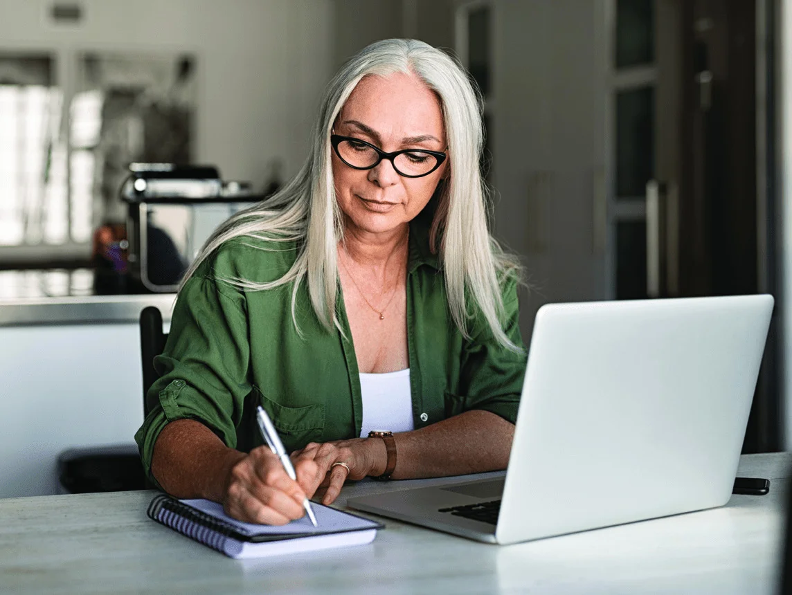 an elderly woman writes in a spiral notebook while looking at laptop